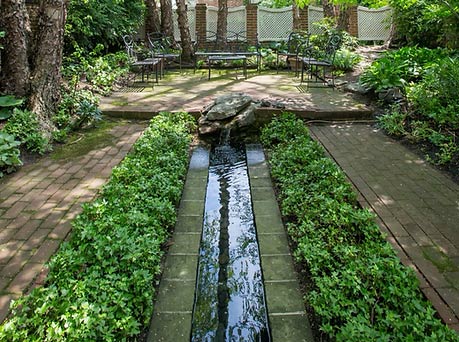 Georgetown Garden Club - Friends of Rose Park Community Partner - image of a water feature with ivy growing on both sides with brick walkways on either side and chairs on a brick patio in a shady yard